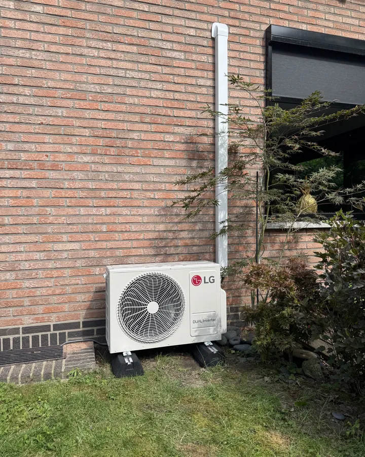 LG airco buitenunit op voetblok in de tuin tegen een bakstenen gevel