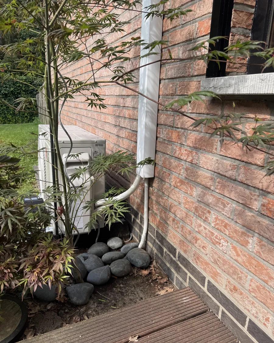 Airco buitenunit weggewerkt tussen de beplanting naast een bakstenen gevel, met de leidingkoker in de kleur van de muur